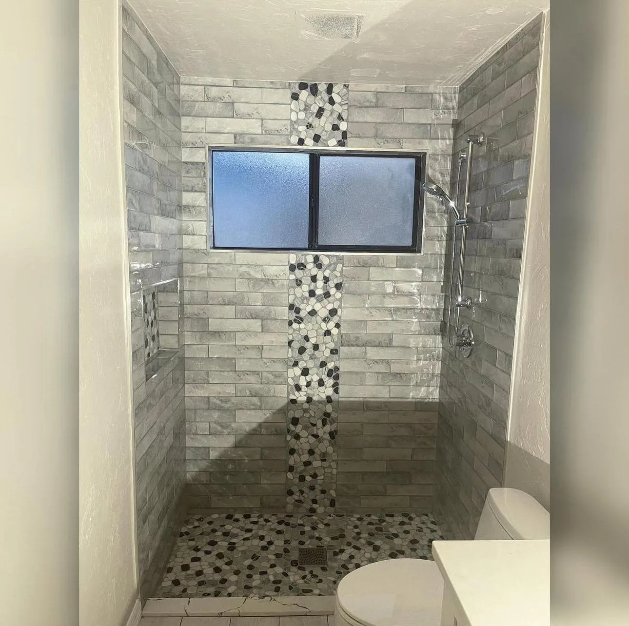 Tiled shower with gray plank tile, pebble mosaic stripe, niche and matching pebble floor in Lakewood Ranch, FL