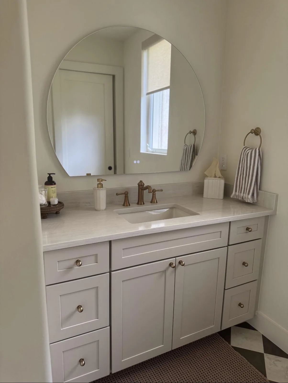 Bathroom vanity with greige shaker cabinets, stone top, brass faucet and round frameless mirror in Lakewood Ranch, FL