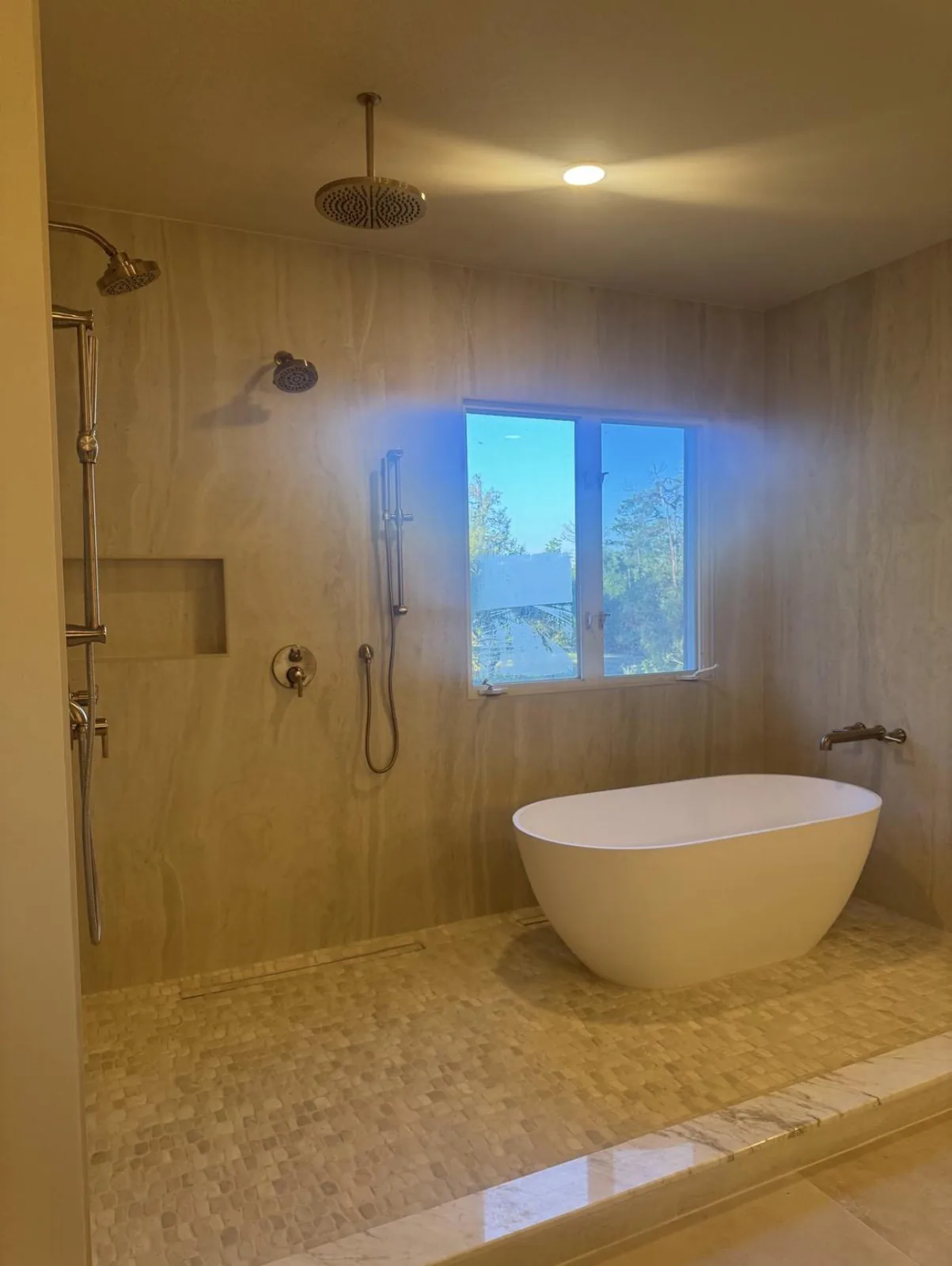 Freestanding soaking tub beside open walk-in shower with travertine-look slab walls and square mosaic floor in Lakewood Ranch, FL