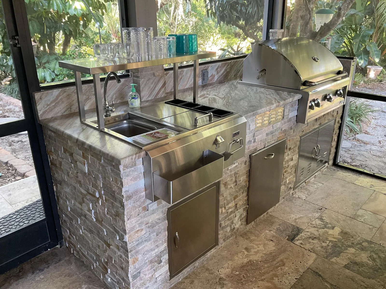 Outdoor kitchen with stacked stone base, natural stone counter, stainless grill, sink and doors in Lakewood Ranch, FL